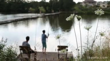 模糊的十几岁的儿子和父亲钓鱼杆钓鱼在木制码头在宁静的池塘前景野花摇曳